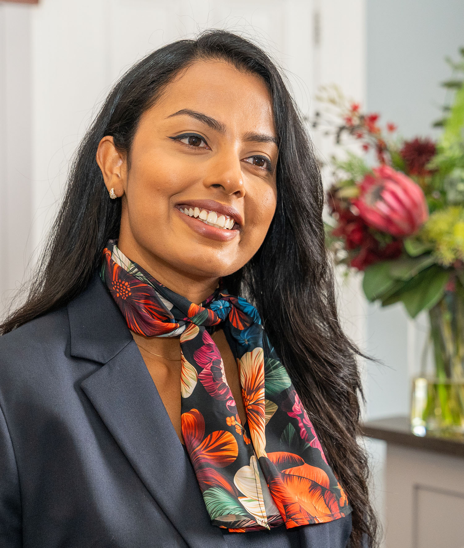 A woman with long brown hair wearing a black blazer and floral neck tie smiling at the camera