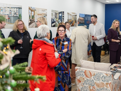  group of people gathered in a modern indoor setting, engaging in conversation during a social or networking event. The room features a blue accent wall, framed interior design photographs on the white wall, and a patterned armchair in the foreground. A decorated Christmas tree is partially visible on the left, and some attendees are holding drinks.