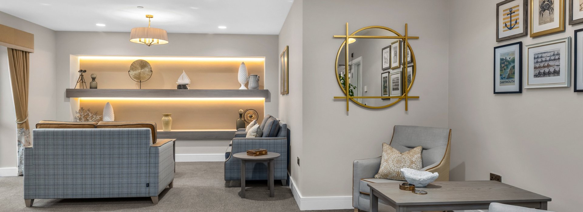 Elegant care home lounge with soft neutral décor, featuring plaid armchairs, a sofa, warm backlit shelving with decorative ornaments, a large round gold-framed mirror and framed artwork on the wall.